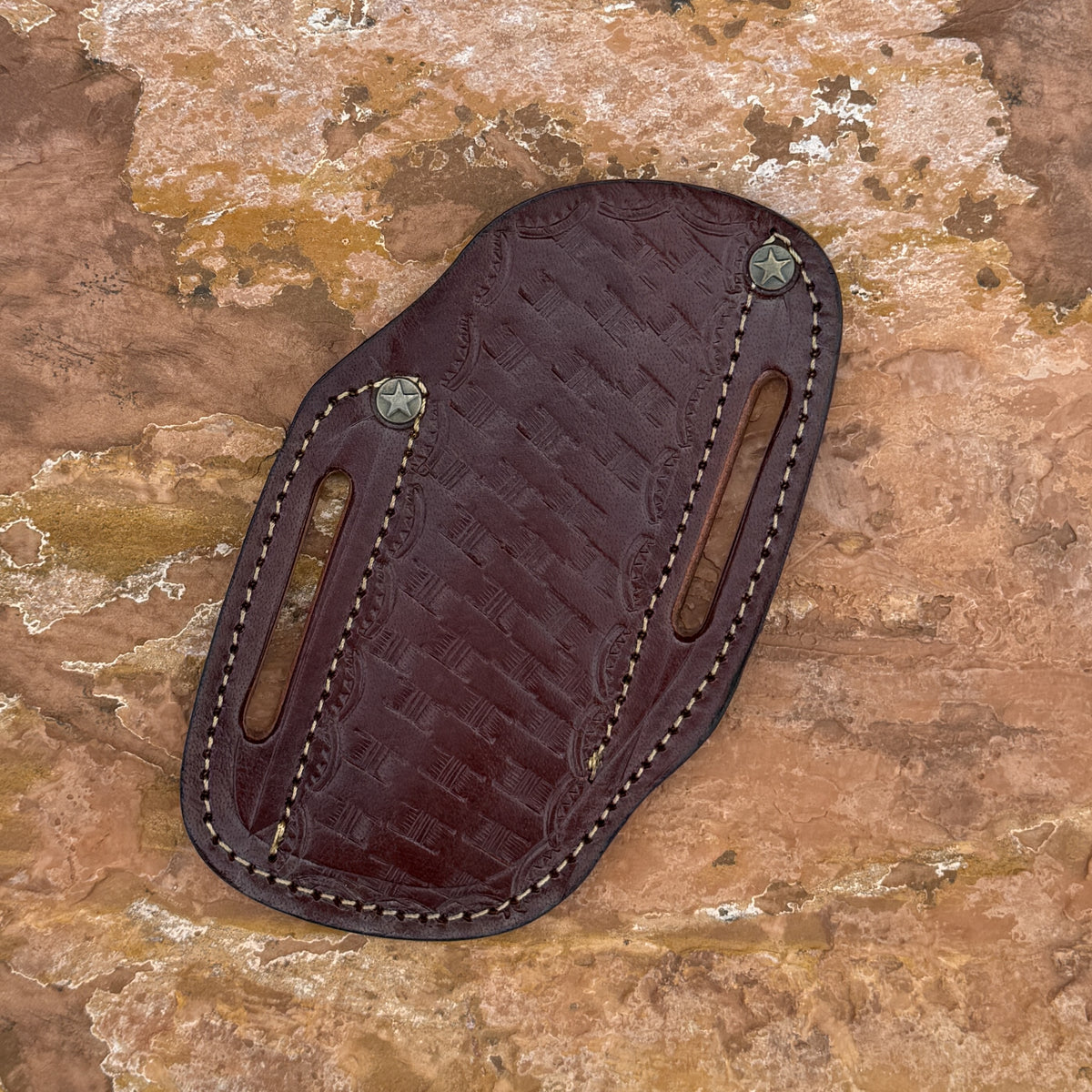 A dark brown embossed leather holster with light tan stitching and metal rivets, laid flat on a textured brown rock surface.